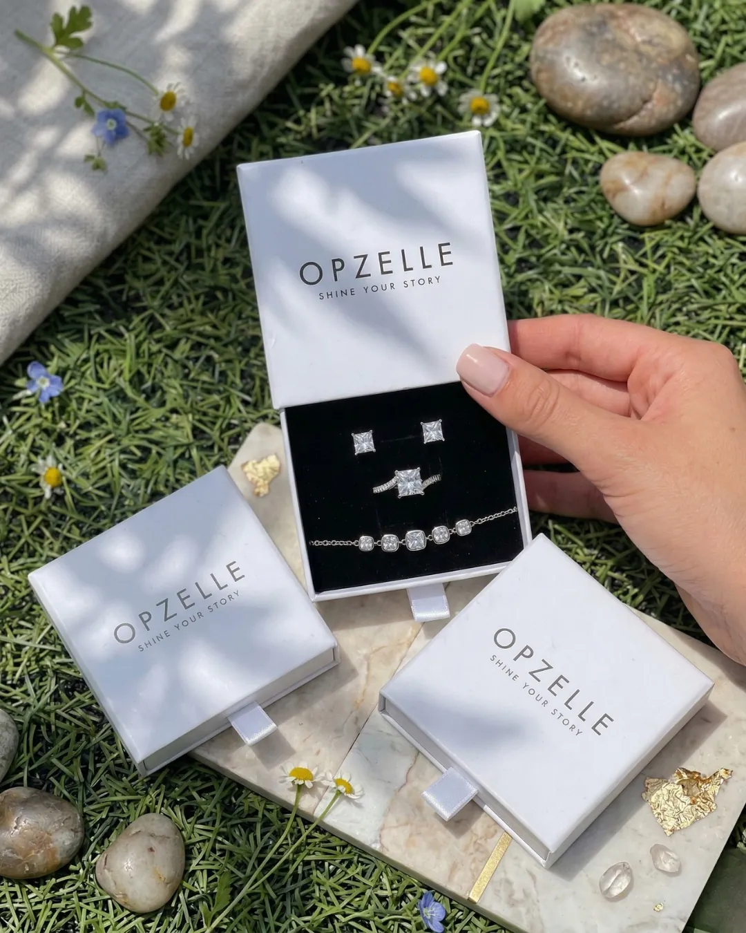 יד פותחת קופסת מגירה של OPZELLE עם עגילים, טבעת וצמיד על קטיפה שחורה